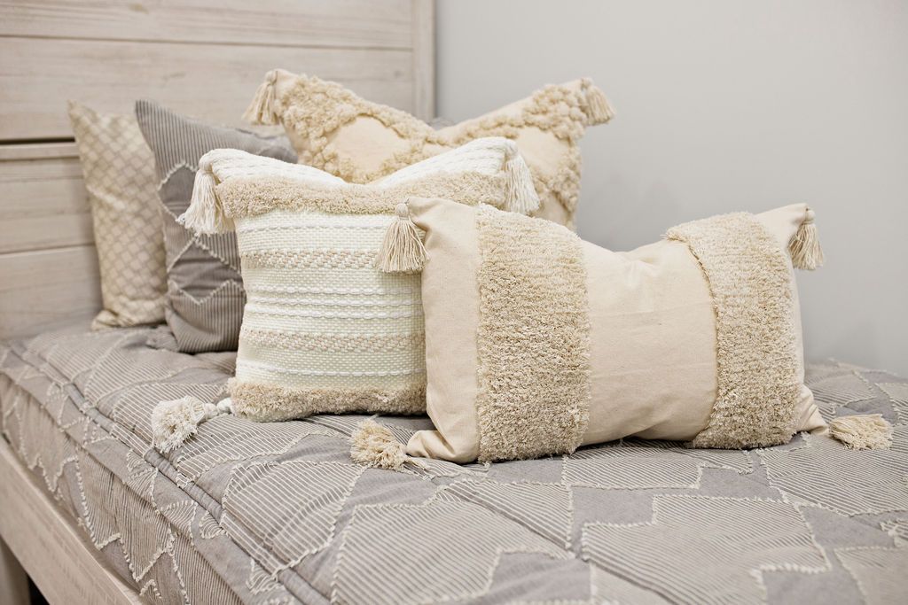 Taupe bedding with textured zig zag design dark creamy textured euro, a cream and tan woven textured pillow and a textured dark creamy lumbar with tassels with a cream knitted chenille throw with pom poms along the edge