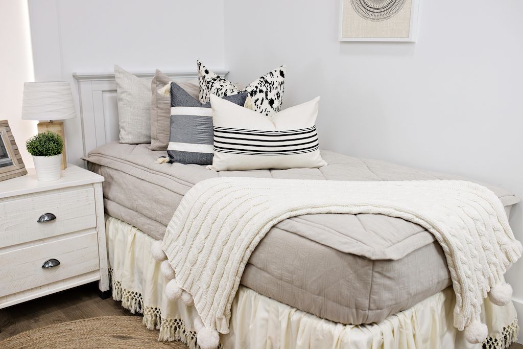 White twin size bed with beige textured bedding, a white and black patterned euro pillow, a medium black and cream textured pillow, a cream and black striped lumbar pillow, a cream bed skirt with tassels and a cream cable-knit throw at the foot of the bed.