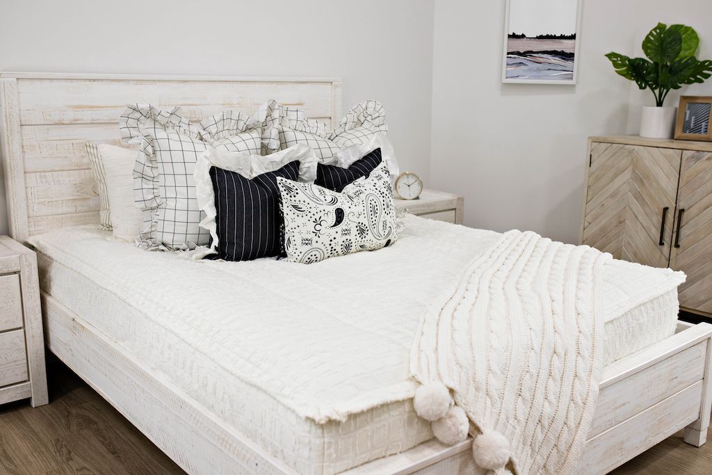 queen bed with cream textured bedding and cream and black grid euro with ruffle along the edge, charcoal striped pillow with white ruffle along the edge, cream lumbar with charcoal paisley print, cream knitted chenille blanket with pom poms