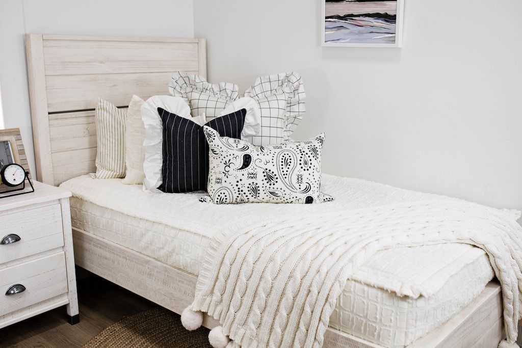 twin bed with cream textured bedding and cream and black grid euro with ruffle along the edge, charcoal striped pillow with white ruffle along the edge, cream lumbar with charcoal paisley print, cream knitted chenille blanket with pom poms