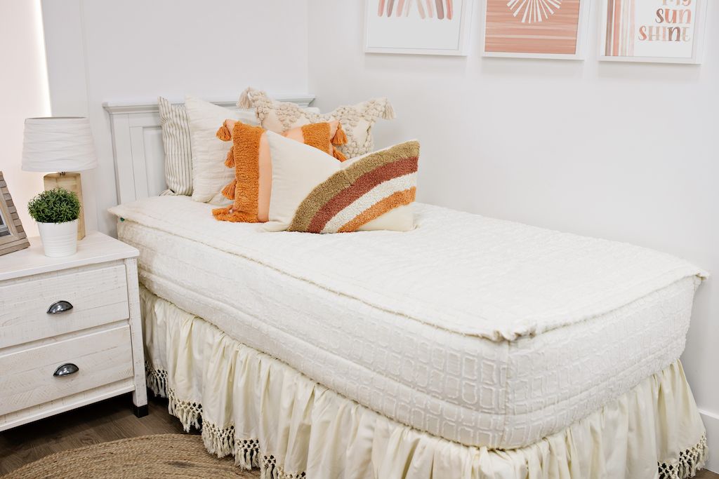 White twin size bed with cream textured bedding, a cream textured euro pillow, a medium peach textured pillow, a boho rainbow lumbar pillow and a cream bed skirt with tassels.