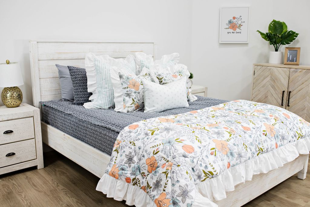 White queen bed with gray and white textured bedding, white and blue striped pillows, multicolored floral pillows, a gray and white lumbar pillow and a multicolored floral blanket.  