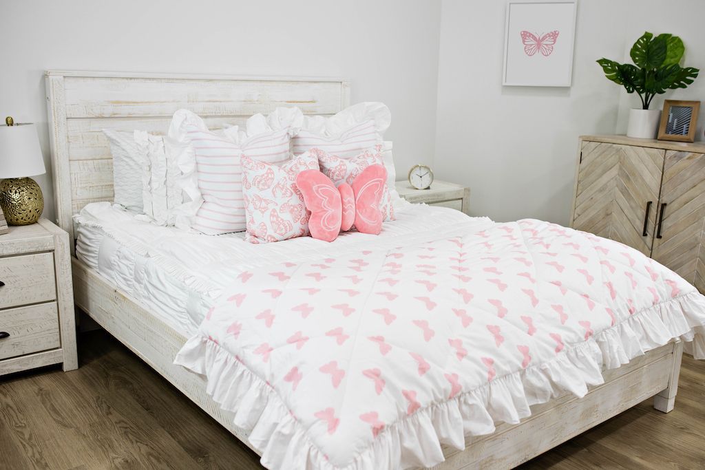 White queen bed with white textured bedding, white and pink striped pillows, pink and white butterfly pillows, a pink butterfly shaped pillow and a white and pink butterfly blanket. 