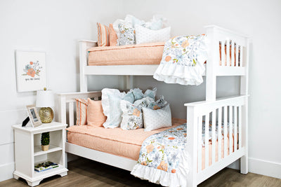 White bunk beds with peach textured bedding, white and blue striped pillows, multicolored floral pillows, a gray and white lumbar pillow and a multicolored floral blanket.  