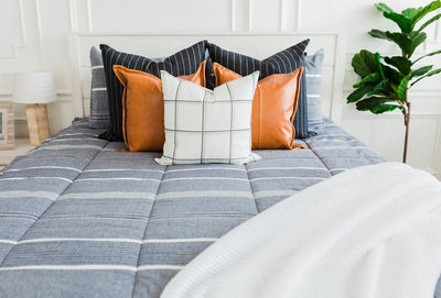 Deep navy woven stripe bedding with deep navy striped euros, faux leather pillows, white and black grid pillow, and white textured blanket with braided tassels