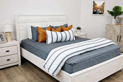 White queen size bed with gray bedding, medium brown leather pillows, a white and gray striped lumbar pillow and a white and gray striped blanket.  