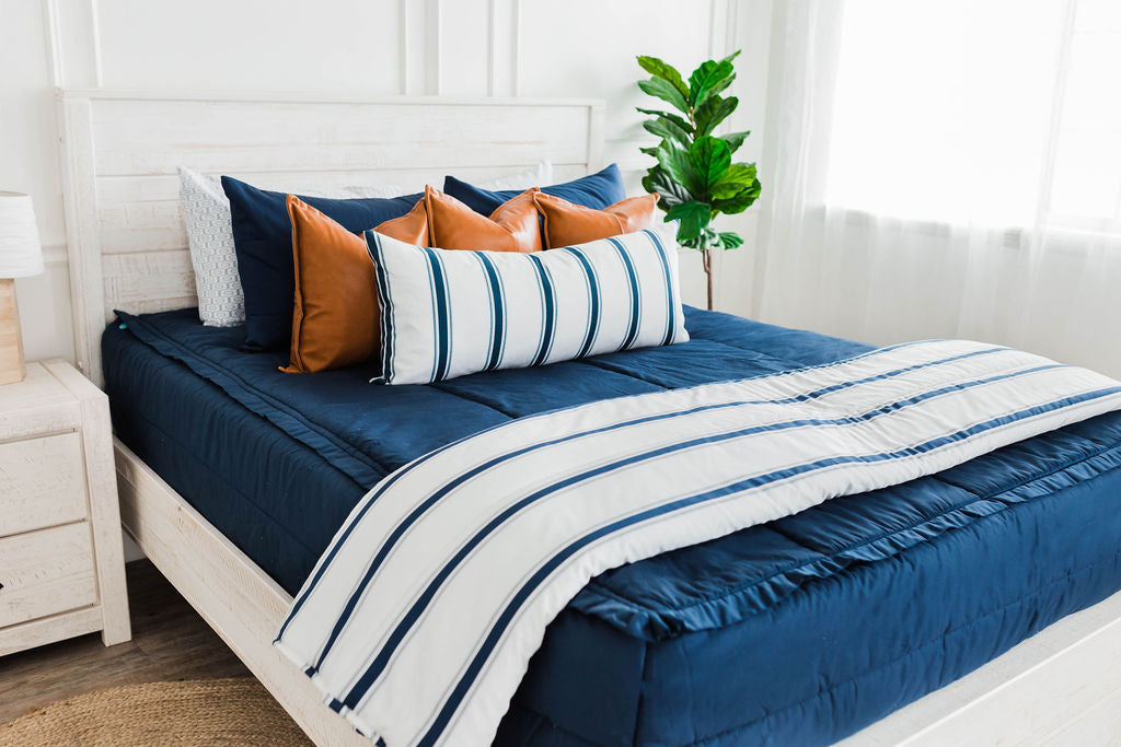 Queen bed with Navy blue zipper bedding with faux leather pillows, white XL lumbar with navy vertical stripes, and white blanket with vertical navy stripes at the foot of the bed