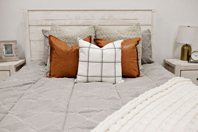 queen bed with Taupe bedding with textured zig zag design, dusty tan faux fur euro, faux leather pillow, white pillow with dark gray grid pattern, and cream knitted chenille throw with pom poms