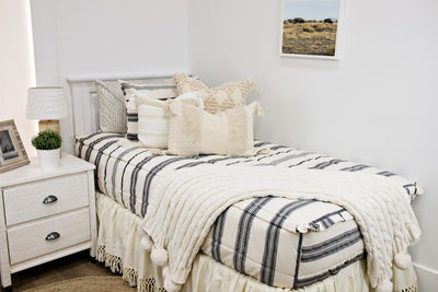 White twin size bed with cream and black woven striped bedding, a cream textured euro pillow, a medium cream textured pillow, a cream textured lumbar pillow, a cream bed skirt with tassels and a cream cable-knit throw at the foot of the bed.