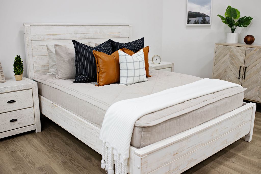 queen bed with beige textured bedding, a navy gray and cream striped pillow, a white and black plaid pillow, a brown leather pillow and a white throw with tassels.