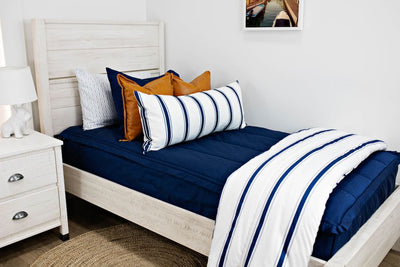 White twin size bed with navy blue bedding, medium brown leather pillows, a white and navy blue striped lumbar pillow and a white and navy blue striped blanket.  