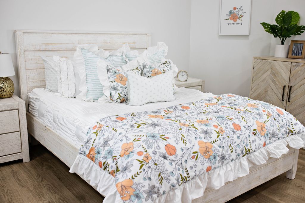 White queen bed with white textured bedding, white and blue striped pillows, multicolored floral pillows, a gray and white lumbar pillow and a multicolored floral blanket.  