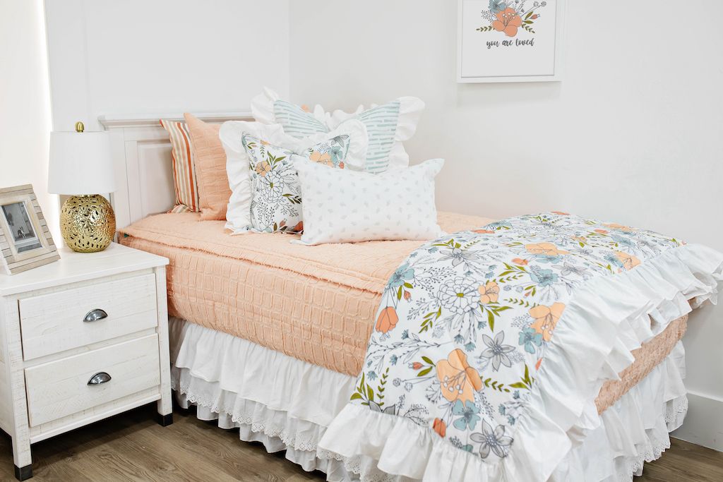 White twin bed with peach textured bedding, a white and blue striped pillow, a multicolored floral pillow, a gray and white lumbar pillow, a white ruffled bed skirt and a multicolored floral blanket.  