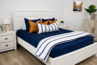 White queen size bed with navy blue bedding, medium brown leather pillows, a white and navy blue striped lumbar pillow and a white and navy blue striped blanket.  