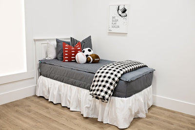 White twin size bed with gray bedding, a black and white striped euro pillow, a medium red and white dash pillow, sport accessory pillows, a white bed skirt and a black and white buffalo checked blanket at the foot of the bed.