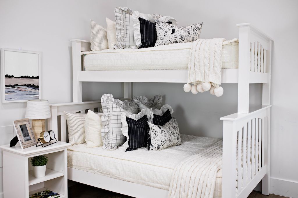 bunk bed with cream textured bedding and cream and black grid euro with ruffle along the edge, charcoal striped pillow with white ruffle along the edge, cream lumbar with charcoal paisley print, cream knitted chenille blanket with pom poms
