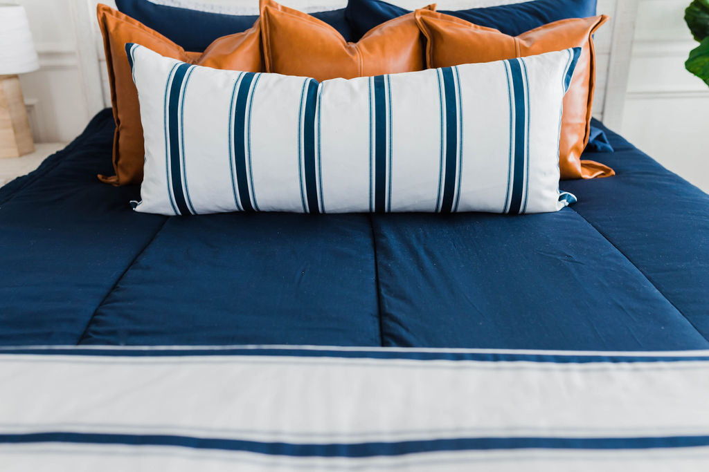 Navy blue zipper bedding with faux leather pillows, white XL lumbar with navy vertical stripes, and white blanket with horizontal navy stripes at the foot of the bed