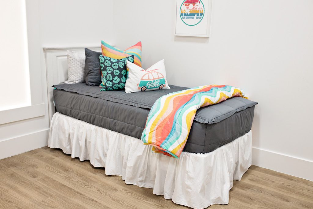 White twin size bed with gray bedding, a multicolored euro pillow, a medium black and green leaf pillow, a white surf bus lumbar pillow, a white bed skirt and a multicolored blanket at the foot of the bed.