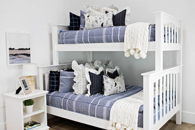 bunk bed with blue striped bedding and cream and black grid euro with ruffle along the edge, charcoal striped pillow with white ruffle along the edge, cream lumbar with charcoal paisley print, cream knitted chenille blanket with pom poms