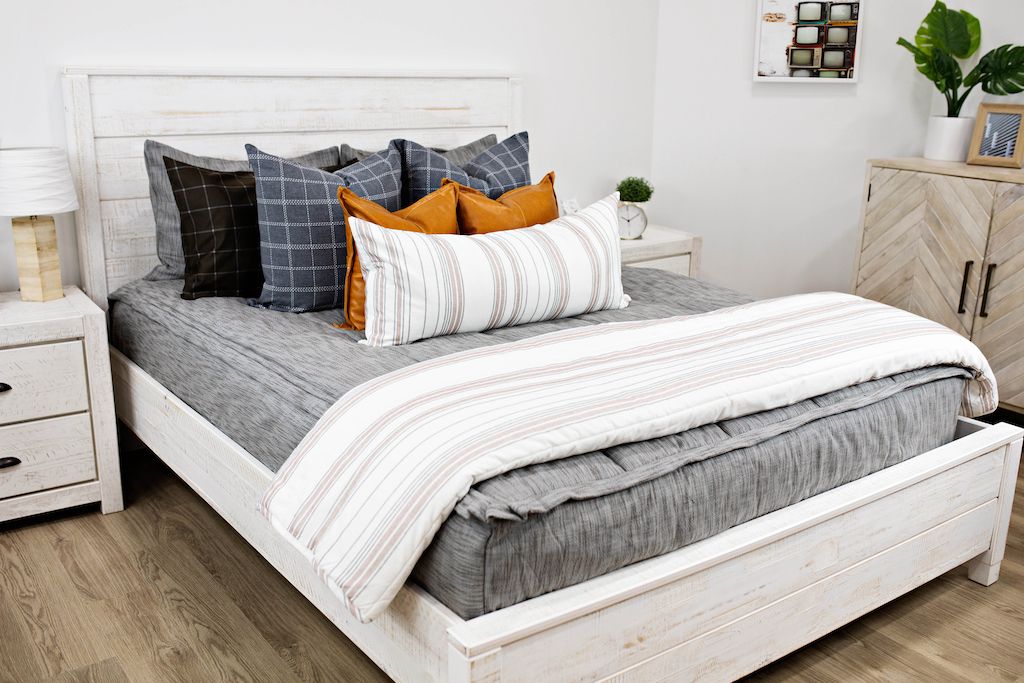 White queen size bed with gray and brown woven bedding, blue and gray plaid euro pillows, medium leather brown pillows, a cream and cognac striped lumbar pillow and a cream and cognac striped blanket.  