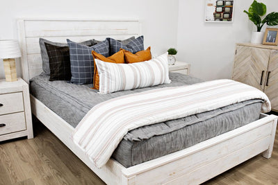 White queen size bed with gray and brown woven bedding, blue and gray plaid euro pillows, medium leather brown pillows, a cream and cognac striped lumbar pillow and a cream and cognac striped blanket.  