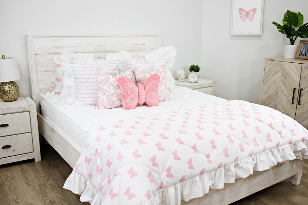 White queen bed with white textured bedding, white and pink striped pillows, white and pink butterfly pillows, a pink butterfly shaped pillow and a white and pink butterfly blanket.  
