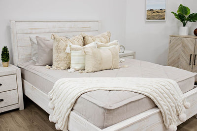 queen bed with tan textured bedding and dark creamy textured euro, a cream and tan woven textured pillow and a textured dark creamy lumbar with tassels with a cream knitted chenille throw pom poms along the edge