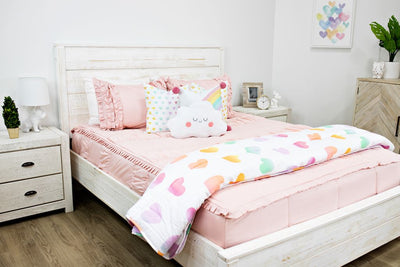 White queen size bed with blush pink ruffled bedding, medium white and rainbow heart pillows, a medium rainbow pillow, a white cloud pillow and a white and rainbow heart blanket. 