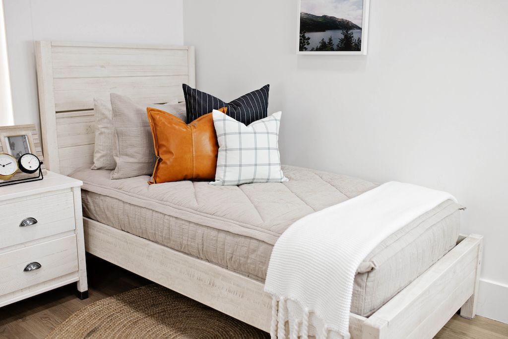 twin bed with tan textured bedding and deep navy striped euros, faux leather pillows, white and black grid pillow, and white textured blanket with braided tassels