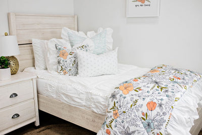 White twin size bed with white textured bedding, a white and blue striped pillow, a multicolored floral pillow, a gray and white lumbar pillow and a multicolored floral blanket.