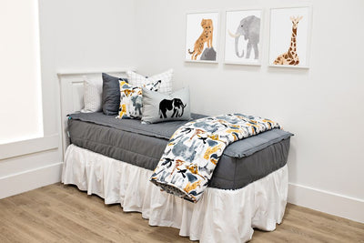 White twin size bed with gray bedding,  a white and black grid euro pillow, a medium white jungle animal pillow, a gray lumbar pillow with an elephant silhouette, a white bed skirt and a white blanket with jungle animals at the foot of the bed.