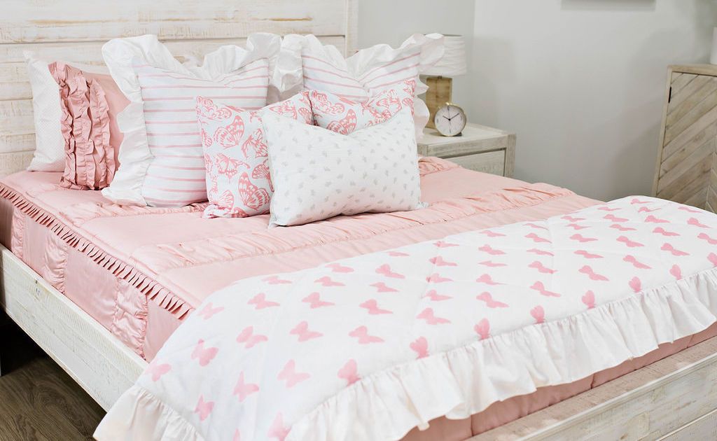 White queen bed frame with pink textured bedding decorated with white and pink striped euro pillows with ruffle along the edge, white medium pillows with pink butterfly design, a white lumbar pillow with small gray floral design and a blanket with pink butterfly design with ruffle along the edge.