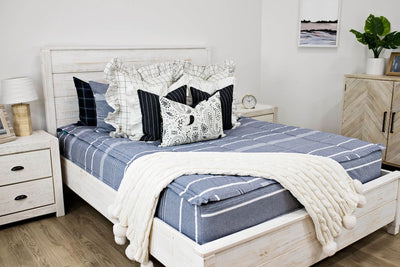 queen bed with blue striped bedding and cream and black grid euro with ruffle along the edge, charcoal striped pillow with white ruffle along the edge, cream lumbar with charcoal paisley print, cream knitted chenille blanket with pom poms