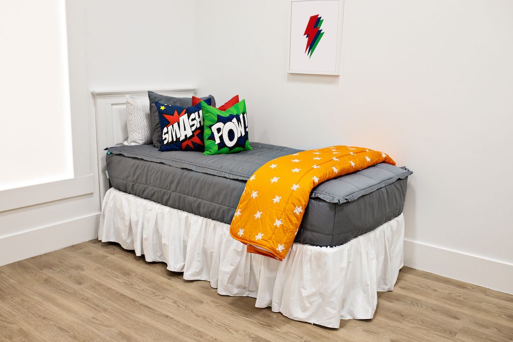 White twin size bed with gray bedding, a medium red pillow, a medium blue pillow, a medium green pillow, a white bed skirt, and an orange and white turtle blanket at the foot of the bed. 