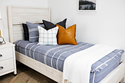 twin bed with blue and navy striped bedding and deep navy striped euros, faux leather pillows, white and black grid pillow, and white textured blanket with braided tassels