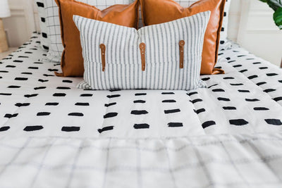 Close up of white bedding with black dashed lines and white and black grid euro, faux leather pillows, white and black striped lumbar with faux lather buckles and white and black grid pattered blanket