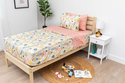A twin bed made with pink, yellow, and blue floral kids zipper bedding, a nightstand beside it with books on the shelf and toys scattered on the floor.