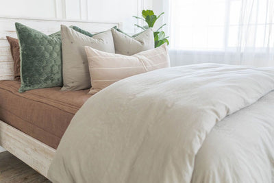 A brightly lit bedroom with a bed made with brown zipper bedding and a slate gray zipper duvet folded half way down the bed and adorned with neutral and green velvet pillows.