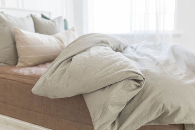 A brightly lit bedroom with a bed made with brown zipper bedding and a slate gray zipper duvet partially folded half way down the bed and adorned with neutral pillows.