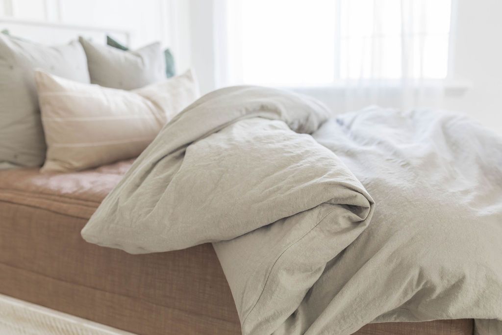 A brightly lit bedroom with a bed made with brown zipper bedding and a slate gray zipper duvet partially folded half way down the bed and adorned with neutral pillows.