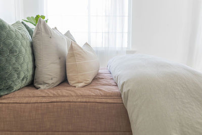 A side view of a bed made with brown zipper bedding and a slate gray zipper duvet folded half way down the bed and adorned with neutral and green velvet pillows.