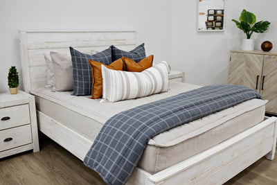 A queen bed made with light tan zipper bedding and neutral gray, leather, and white pattern pillows and matching blanket.