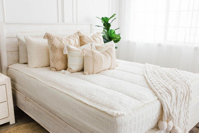 queen bed with Cream bedding with textured rectangle design and dark creamy textured euro, a cream and tan woven textured pillow and a textured dark creamy lumbar with tassels with an off white braided throw with pom poms along the edge