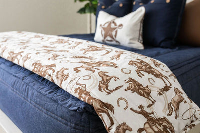A close up of a bed made with denim blue zipper bedding and neutral pillows and a white blanket with a cowboy and horseshoe design at the end of the bed.