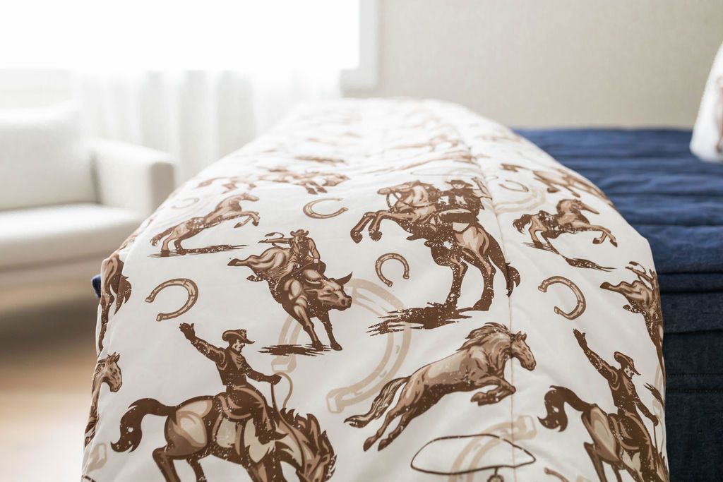 A close up side view of a bed made with denim blue zipper bedding with a cowboy and horseshoe printed blanket at the end of the bed.