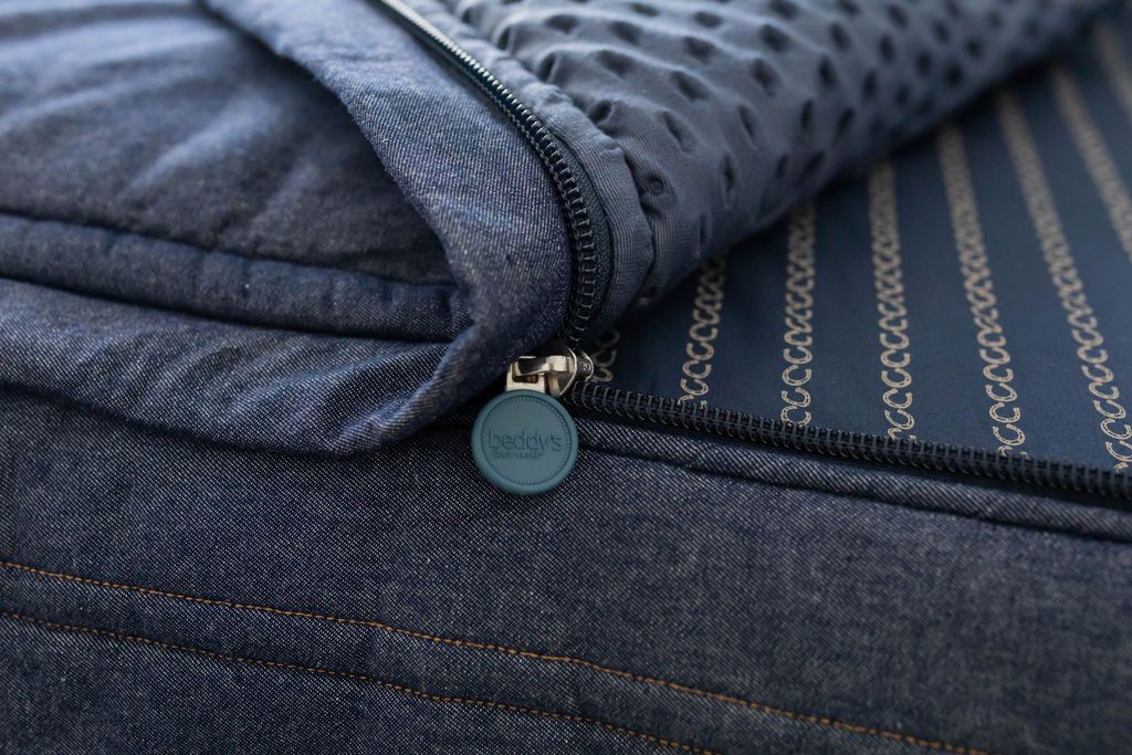 A close up view of a bed made with dark denim blue zipper bedding with the bed slightly unzipped to reveal the minky comforter and horseshoe striped pattern bottom cotton lining.