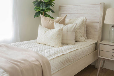 A twin bed made with cream embroidered zipper bedding and adorned with neutral patterned pillows and a throw tan blanket.