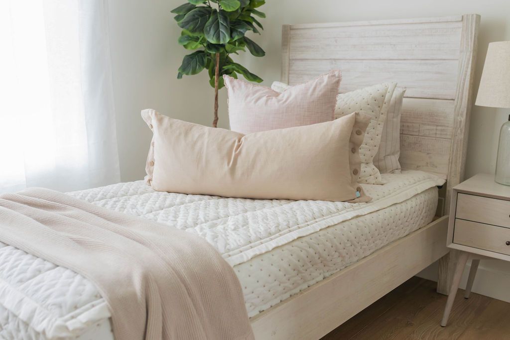 A twin bed made with cream embroidered zipper bedding and adorned with neutral and light pink pillows and a throw tan blanket.