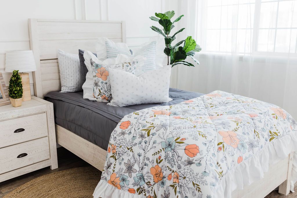 twin bed with Gray zipper bedding with white and blue striped euro with ruffle along the edge, floral printed pillow, white lumbar with small gray floral design and floral printed blanket with white ruffle along the edge