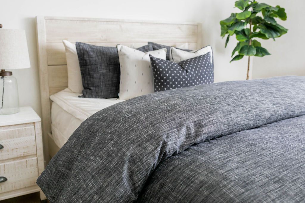 A brightly lit bedroom with a bed made with white zipper bedding and a dark gray zipper duvet folded half way down the bed and adorned with neutral black and white pillows.